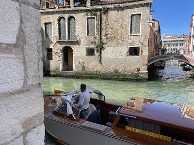 Living the Life In Venice Italy Why Extended Travel is the Secret to a More Fulfilling Retirement. A wooden taxi boat navigates a narrow Venice canal, with a man standing at the helm. Weathered stone and stucco buildings line the canal, one bearing graffiti and street art near a boarded window, with a stone archway and small bridge visible in the background."