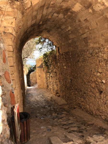 Meandering Your Way Through Retirement Why extended travel is the secret to a more fulfilling retirement. A narrow stone passageway curves through an arched tunnel built into a fortress wall, with dappled sunlight filtering through greenery at the far end. A glimpse of blue sea is visible through the opening, and a small bin and leaning signs sit near the tunnel's entrance."