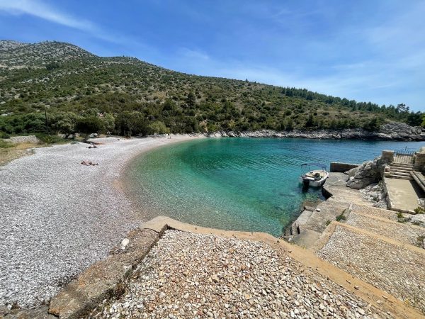 Dubovica Beach Hvar A secluded pebble beach at Dubovica, Hvar, Croatia, curving around a bay of clear turquoise water that deepens to teal in the center. A small white motorboat is moored near stone steps on the right, with a green, tree-covered hillside rising behind the cove under a bright blue sky.