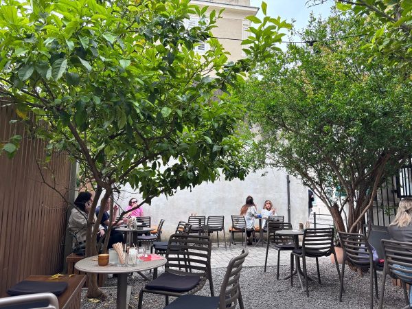 Kava2 Cafe Split Croatia A shaded outdoor café courtyard in Split, Croatia, with round tables and grey chairs set on a gravel and paved floor, canopied by leafy citrus trees. Several people sit chatting at the tables, with a pale stone building visible in the background.