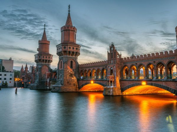 Oberbaum Bridge, Kreuzberg, Berlin Best accommodation in Berlin. The historic Oberbaum Bridge in Berlin at twilight, constructed from red brick with twin turrets and prominent arches over the River Spree, glowing warm yellow from underspanning lights reflected on the water.