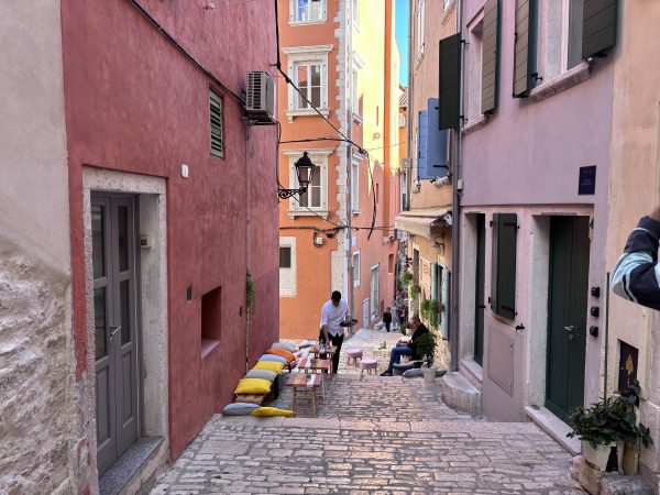 Trevisol Bar Rovinj Istria, Croatia A steep cobblestone alley between colorful pink, orange, and yellow buildings in Rovinj, Istria, with a waiter serving an outdoor café seating area furnished with cushioned benches and small striped tables.