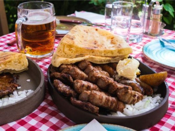 A dark oval dish piled with rows of grilled ćevapi sausages over chopped raw onion, topped with a wedge of grilled flatbread, a scoop of creamy kajmak, and a grilled pepper wedge. The dish sits on a red-and-white checkered tablecloth beside a mug of beer, water glasses, and another plate of grilled flatbread with meat and onions.
