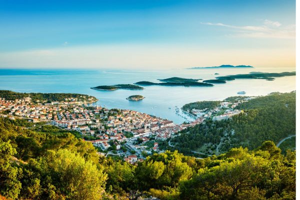 Views from the Castle of Hvar Town on Hvar Island, Croatia Day trips from Split. An elevated scenic view of Hvar town in Croatia, showing red-roofed buildings nestled along a curved harbor of turquoise sea water. Lush green pine trees frame the foreground, while small green islands dot the calm, sunlit blue Adriatic Sea stretching out to the horizon under a clear sky.