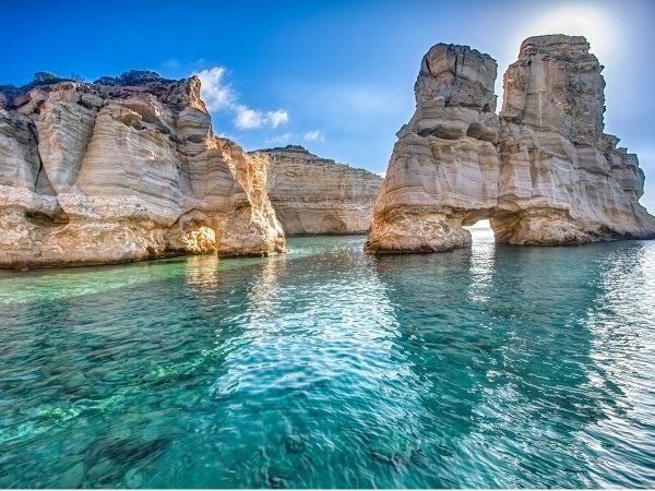 Exploring the Coast of Milos Scenic coastal view with clear turquoise water reflecting sunlight, large beige rock formations with natural arches, under a bright blue sky. Serene and majestic.