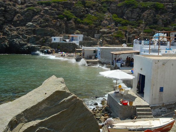 The Ancient Port of Seralia, Sifnos, Greece (1) Coastal scene with white buildings and a terrace overlooking turquoise waters. People enjoy a small beach, waves splash against rocks, evoking a serene, lively atmosphere.