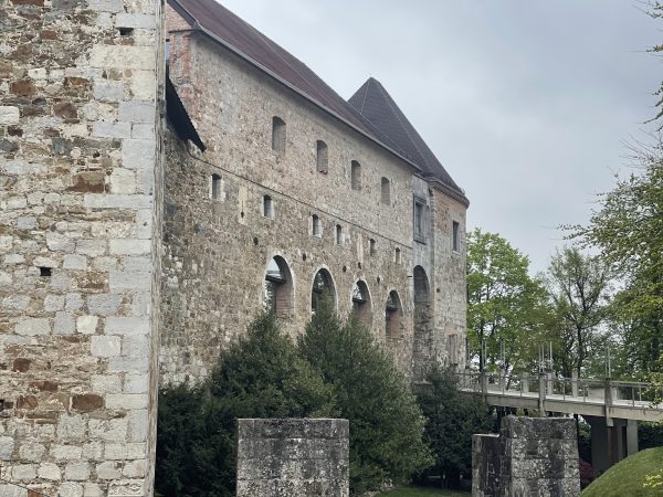 Rustic stone entrance to Ljubljana Castle featuring arched walls, a tall medieval tower, and a drawbridge crossing over a moat. The historic fortress architecture creates a dramatic entrance to one of Ljubljana’s most famous landmarks.