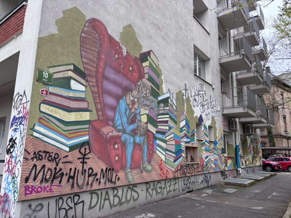 Vibrant street art mural in Ljubljana showing an old man with a long beard wearing a blue suit while reclining on a lounge chair surrounded by towering stacks of books. The detailed artwork reflects the creative and artistic atmosphere found throughout the city’s alternative neighborhoods.