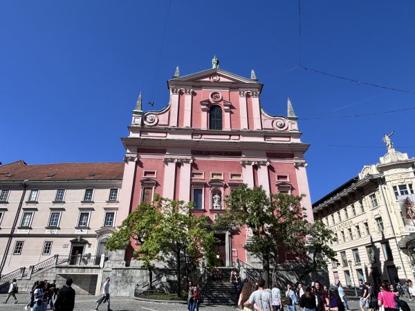 Franciscan Church of the Annunciation in Ljubljana with its salmon pink Baroque facade overlooking Prešeren Square near the Triple Bridge. The historic church is decorated with intricate architectural details and topped with a copper statue of Our Lady of Loretto, standing out as one of the city’s most recognizable landmarks.