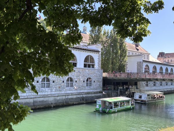 Beautiful Spring Day cruising down the River Ljubljanica. The water is emerald green and the cliccial building behind is white and stone with many arches.