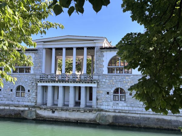 Elegant riverside building in Ljubljana with a light colored facade and large semi circular windows overlooking the river. A classical colonnade lines the street side, creating one of the city's most distinctive architectural landmarks along the waterfront.