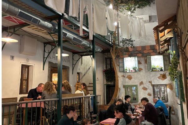 Courtyard dining area at Baščaršija restaurant in Ljubljana designed to resemble an old town square with washing hanging overhead, potted plants, and red checked tablecloths. Guests sit enjoying traditional local meals while live music creates a warm and lively atmosphere.
