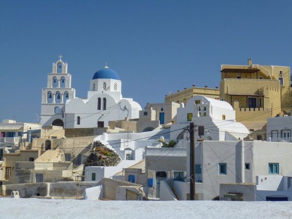 Santorini skyline with whitewashed buildings, blue domed church, and bell tower under a clear blue sky, conveying a serene Mediterranean vibe.