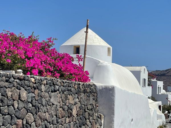 White dome-shaped building with vibrant pink bougainvillea flowers atop a stone wall under a clear blue sky, creating a serene Mediterranean scene.