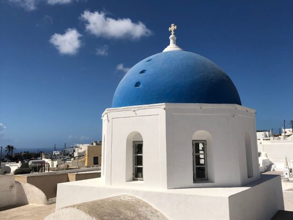 White building with a vivid blue dome and a cross on top, against a clear sky with scattered clouds. The scene is bright and tranquil.