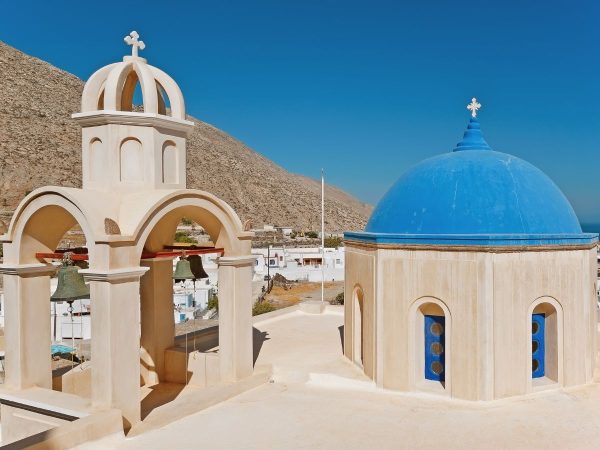 White church with a blue domed roof and arches against a clear blue sky in Santorini. Bells hang under arches, evoking a calm, serene atmosphere.
