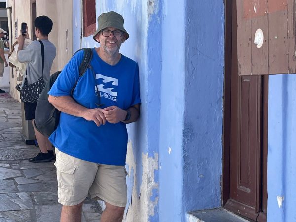A smiling man in a blue t-shirt and green hat leans against a blue wall in a narrow street. He holds a camera and a bag, exuding a relaxed, joyful vibe.