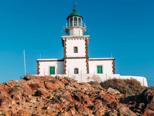 White lighthouse with a green dome sits atop a rocky hill under a clear blue sky. The building's edges are accented with brown bricks, conveying tranquility.