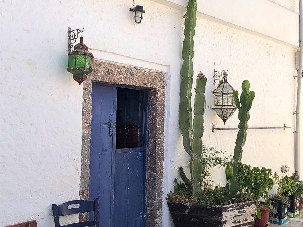 Outdoor scene with a rustic blue door framed in stone, beside a tall potted cactus. Two green hanging lanterns adorn the white textured wall.