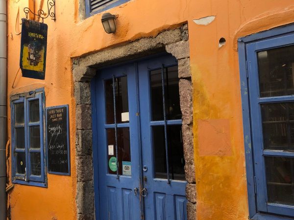 Vibrant orange facade with rustic blue doors and windows. A sign for "Yira Taverna Jazz Bar" evokes a cozy, inviting atmosphere.
