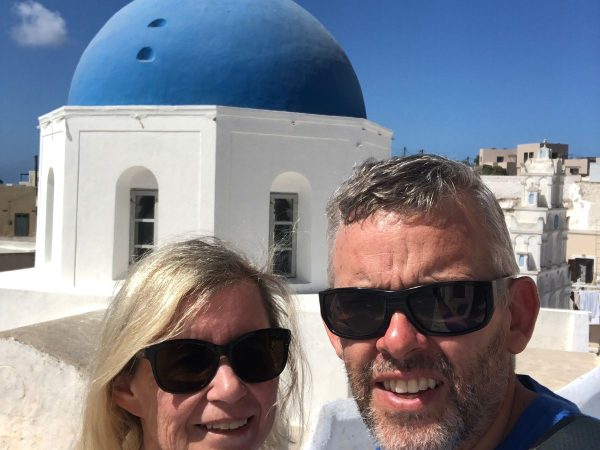 A couple wearing sunglasses smiles in front of a white building with a prominent blue dome, set against a clear blue sky in a sunny Mediterranean setting.