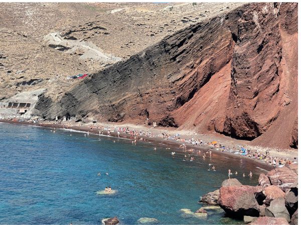 A scenic beach with red cliffs and clear blue water. People are sunbathing and swimming. The atmosphere is lively and relaxed, with rugged, natural terrain.