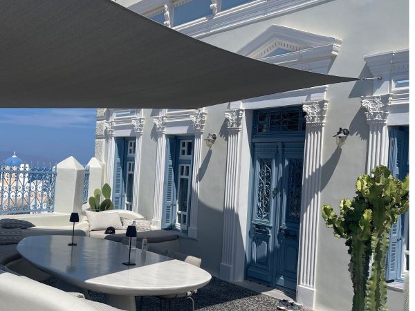 A Mediterranean-style terrace with white walls, blue doors, and windows. A shade sail covers a marble table and cozy seating. A cactus and sea view add a serene vibe.