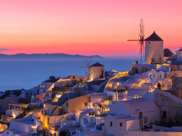 Sunset over Santorini, Greece, with white buildings and a windmill glowing under pink-orange skies. The sea and distant islands create a serene backdrop.