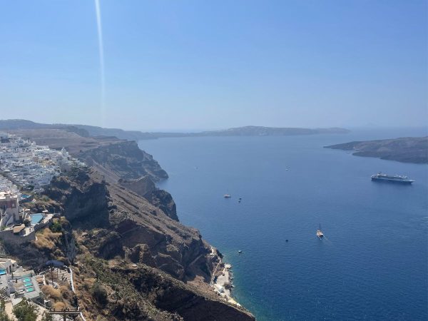 A serene coastal view of Santorini with white buildings atop rugged cliffs, overlooking a deep blue sea. A cruise ship and boats dot the calm water.