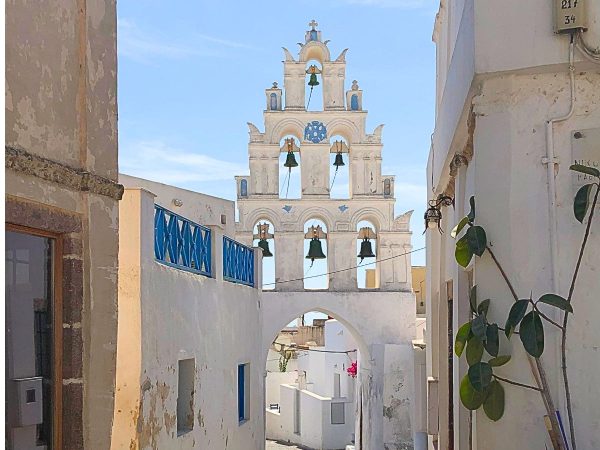 A narrow alley with white, sunlit walls leads to an intricate bell tower featuring multiple arches and blue accents. Clear blue sky enhances the serene atmosphere.