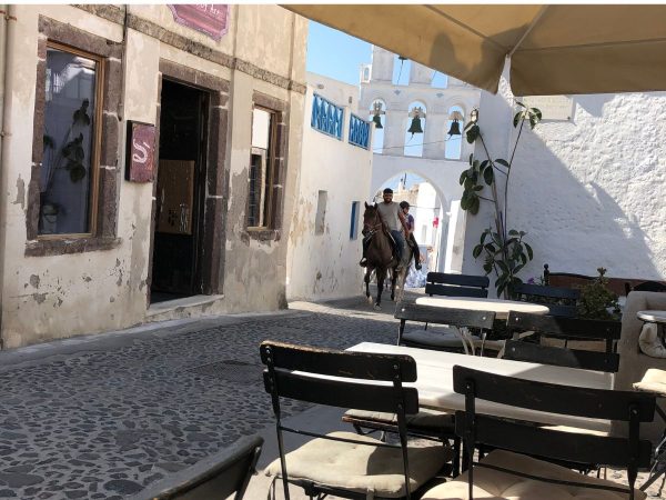 A sunny, narrow cobblestone street in a quaint village, with two horses carrying people. Empty cafe seats in the foreground evoke a tranquil ambiance.