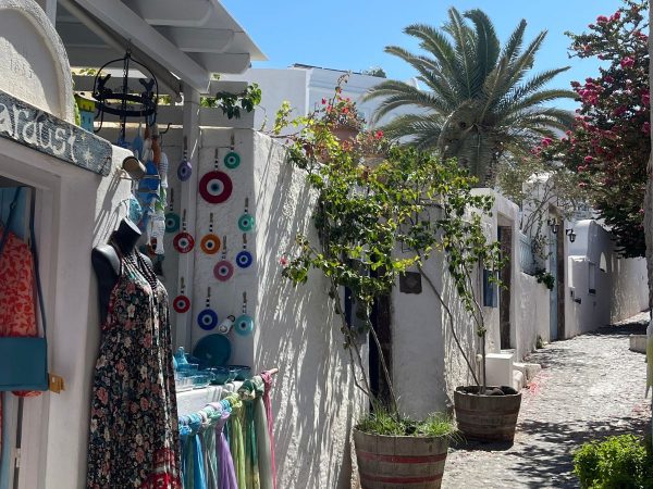 Charming cobblestone street lined with white walls, vibrant hanging trinkets, and colorful sarongs. A mannequin displays a floral dress. Palm trees and bougainvillea add a Mediterranean feel under a clear blue sky.