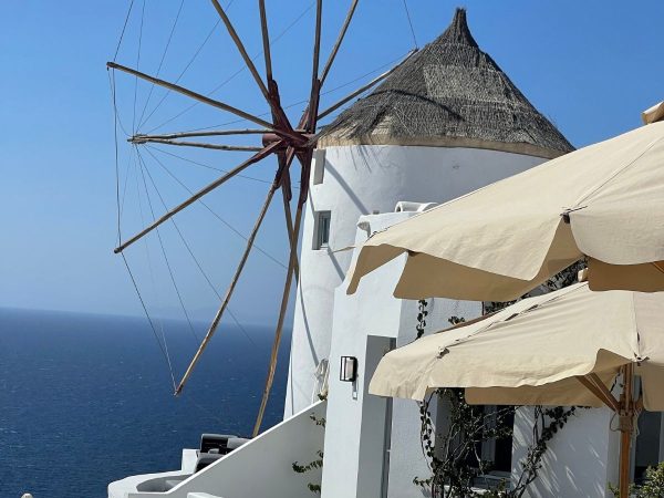 A white windmill with a thatched roof overlooks the deep blue ocean under a clear sky. Beige umbrellas add a relaxed, sunny atmosphere.