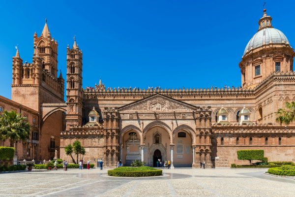 Palermo Cathedral is one of the most architecturally extraordinary buildings in the entire Mediterranean — a UNESCO World Heritage listed structure that was successively a Norman cathedral. Palermo Cathedral rises from its marble courtyard in extraordinary architectural complexity — neatly clipped hedges frame the formal approach while the limestone facade above tells the story of a thousand years of Sicilian history in stone, soaring Catalan Gothic porticos and pointed arches sitting alongside intricate Moorish geometric patterns and interlacing Norman arches, the original Norman apses still intact at the rear as if anchoring the whole extraordinary building to its earliest origins.