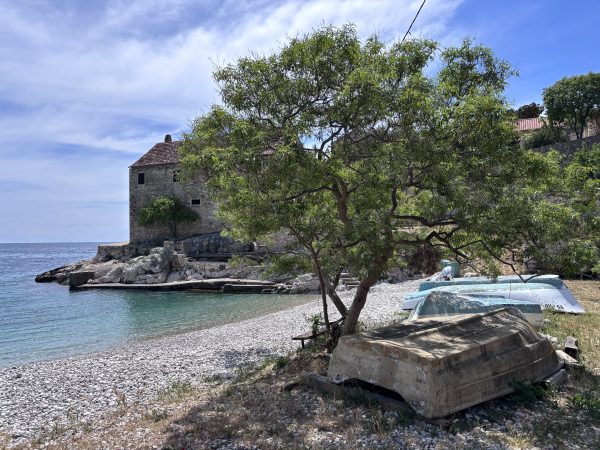 Dubovica Beach Hvar Day Trips from Split where you see a small stone building on a rocky point beside a calm cove, with a leafy tree and old wooden boats resting on the pebble shore.