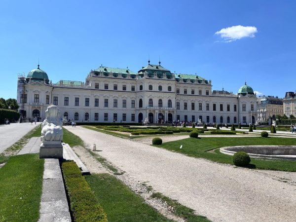 Historic palace with a baroque architectural style, featuring a grand facade with green roofs. The foreground shows manicured gardens and a statue under a clear blue sky.
