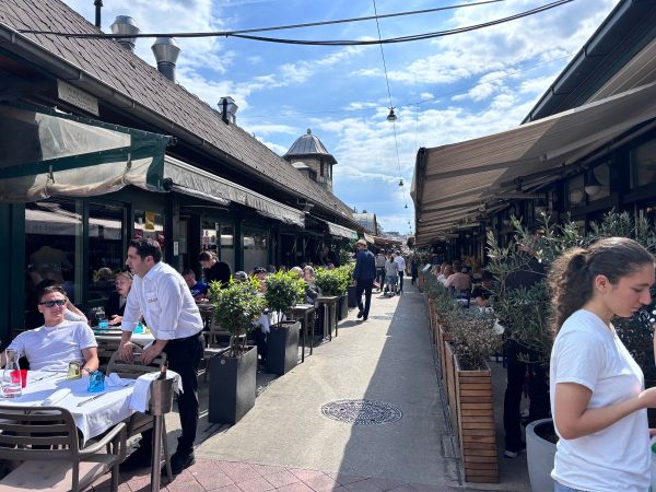 A bustling outdoor cafe scene in a narrow alley, with patrons dining under awnings. A waiter attends to a table. The atmosphere is lively and bright.