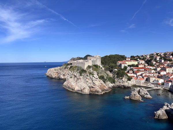 Fort Lovrijenac, a stone fortress perched atop a rocky promontory in Dubrovnik, Croatia, rising above deep blue sea with rocky outcrops jutting from the water below. Terracotta-roofed buildings of the Old Town spread across the hillside to the right, under a clear blue sky with light wispy clouds.