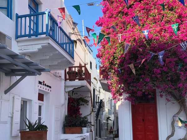 Accommodation in Europe Mykonos Greece A narrow whitewashed alley in Mykonos, Greece, with a bright red door and a blue-shuttered window on one side, and a vibrant magenta bougainvillea plant cascading over a white wall on the other. Colorful triangular flags with religious symbols are strung overhead between the buildings, and potted plants line the base of the walls under a clear blue sky