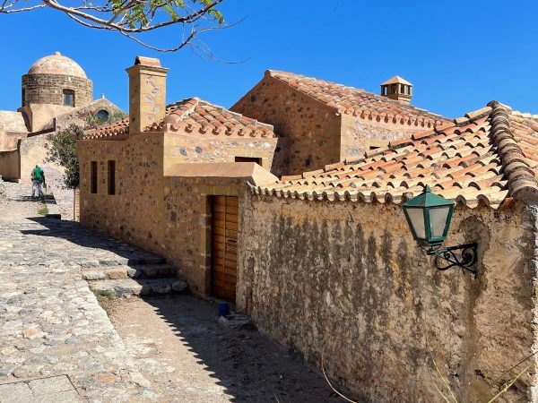 Accomodation In Europe, Monemvasia, Greece A sunny street scene in Monemvasia, Greece, showing stone houses with terracotta tiled roofs and a domed stone building in the background. A green lantern hangs on an iron bracket mounted to a weathered stone wall, a wooden door sits beneath a small chimney, and a person in a green shirt walks along the cobblestone path under a clear blue sky.