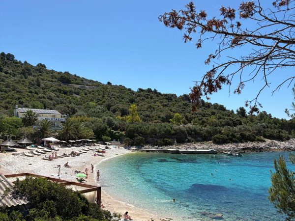 Beach Cove with pebbles on the beach and turquoise blue water.  There are people  on the  beach , sunbeds,  umbrellas and a building. 