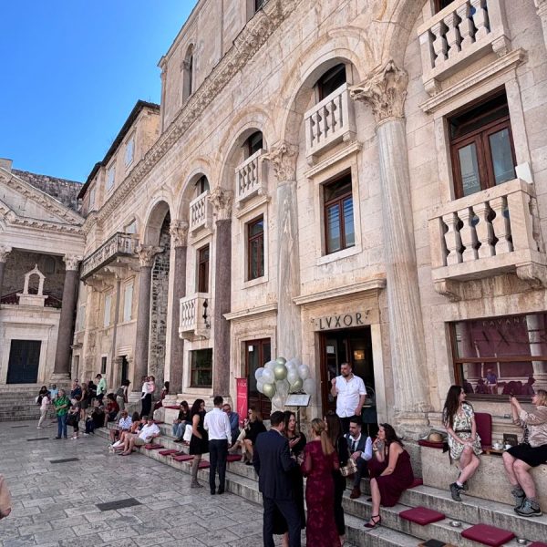 "A sunlit street scene in Split's old town, showing the ornate limestone facade of the Peristyle square with tall Roman columns, arched windows, and stone balconies with balustrades. A sign reading 'LVXOR' marks a bar entrance, with silver balloons tied near the doorway. People in evening wear sit and stand on wide stone steps cushioned with dark red seat pads, socializing in the golden late-afternoon light. A cafe with outdoor seating is visible to the right, and pedestrians walk along the paved street below a bright blue sky."