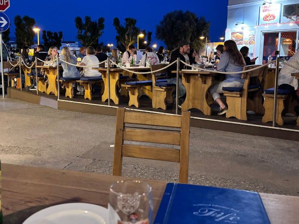Rows of wooden picnic-style tables with blue cushioned benches, filled with diners, set outdoors along a waterfront at dusk under a deep blue sky. A lit sign for a Turkish restaurant, "Lafido," glows in the background, and an empty wooden chair sits in the foreground. One of the amazing restaurants in Split. 