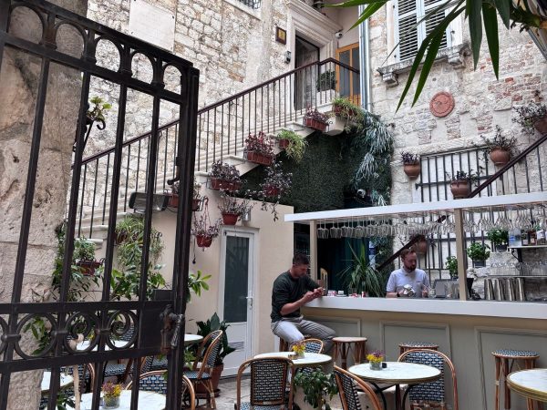 Judita Palace Heritage Hotel Split An open wrought-iron gate leads into a stone courtyard bar with small round marble tables, patterned chairs, hanging potted plants along a staircase, and two men standing at a white outdoor bar counter lined with hanging wine glasses. bar in Split Croatia