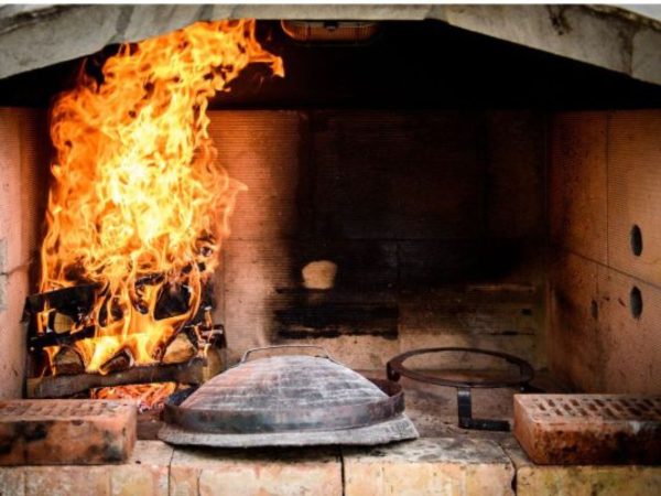 Inside a stone fireplace, tall orange flames burn over a pile of logs on the left side. On the brick hearth in front, a rounded metal peka lid with a handle sits beside a round metal grate, with sooty brick walls and two loose bricks resting nearby.  One of the many restaurants in Split. 