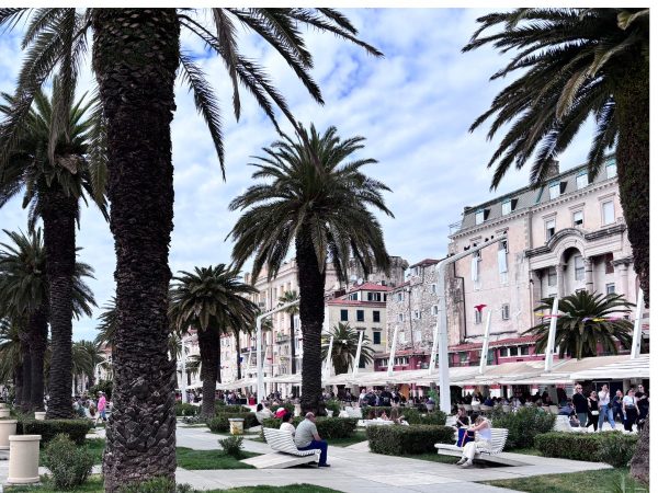A palm-tree-lined waterfront promenade with white benches where people sit and relax, backed by a row of elegant pastel-colored historic buildings under a partly cloudy sky. Colorful flags flutter from lampposts along the walkway. Showcasing the restaurants in Split.