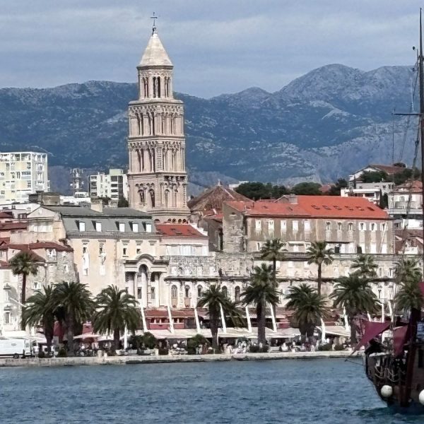 A view of Split's waterfront from across the harbor, dominated by the tall stone bell tower of Saint Domnius Cathedral rising above the old town's terracotta rooftops and historic buildings. A row of palm trees lines the promenade along the water's edge, with outdoor cafe umbrellas visible beneath them. Rugged gray mountains form the backdrop under a partly cloudy sky, and the rigging of a wooden sailing ship is visible in the foreground on the right, with calm blue water in front.