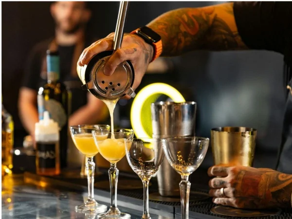 A tattooed bartender strains a cocktail from a metal shaker into a row of coupe glasses, with another bartender working in the background and a glowing yellow neon light behind the bar.
