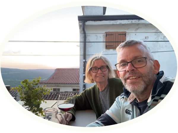 A man and woman smiling for a selfie at an outdoor table during golden hour, with a glass of red wine, olive oil bottle, and small potted plant in the foreground, and a whitewashed building with a wooden shutter and terracotta rooftops behind them overlooking a hazy valley.