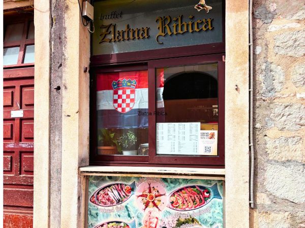 The stone entrance of a restaurant called "Zlatna Ribica," with gold gothic-style lettering above a window. A Croatian flag with its red-and-white checkered crest hangs inside the window next to a printed menu, and a colorful poster below the window advertises seafood dishes like mixed fish, salt sardines, and fried shrimp. One of the Amazing Restaurants in Split. 